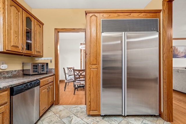 kitchen with a toaster, appliances with stainless steel finishes, brown cabinets, dark stone countertops, and glass insert cabinets