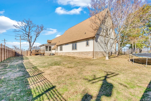 back of property with a fenced backyard, roof with shingles, a trampoline, a deck, and a yard