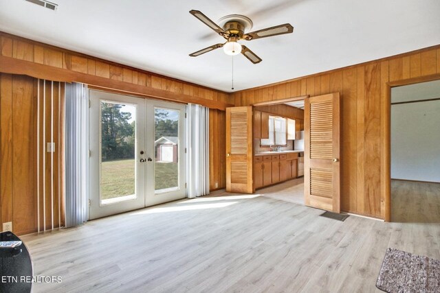 unfurnished living room with a ceiling fan, french doors, wood walls, and light wood finished floors