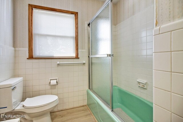 bathroom with a wainscoted wall, shower / bath combination with glass door, toilet, wood finished floors, and wallpapered walls