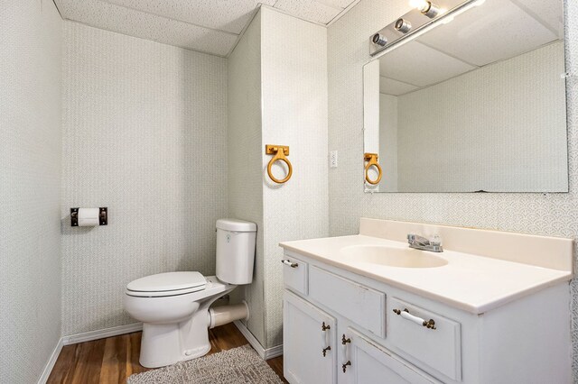 bathroom with toilet, vanity, wood finished floors, a drop ceiling, and wallpapered walls