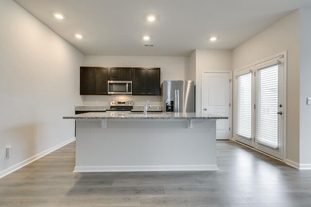 kitchen with stainless steel appliances, light stone counters, an island with sink, and light wood finished floors