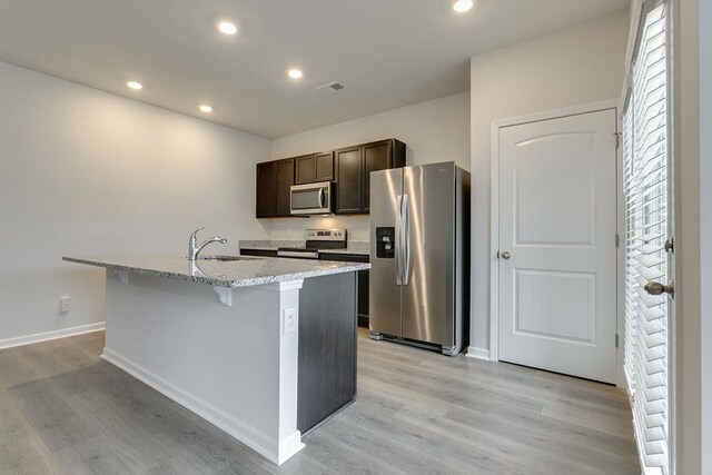 kitchen featuring light stone counters, a kitchen island with sink, dark brown cabinetry, stainless steel appliances, and a sink