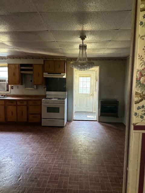 kitchen featuring brick floor, under cabinet range hood, brown cabinetry, gas range gas stove, and decorative light fixtures