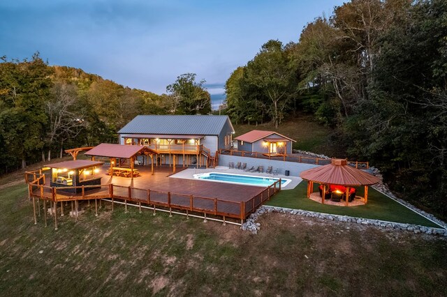 back of house at dusk with a wooden deck, a fenced in pool, metal roof, a gazebo, and a yard