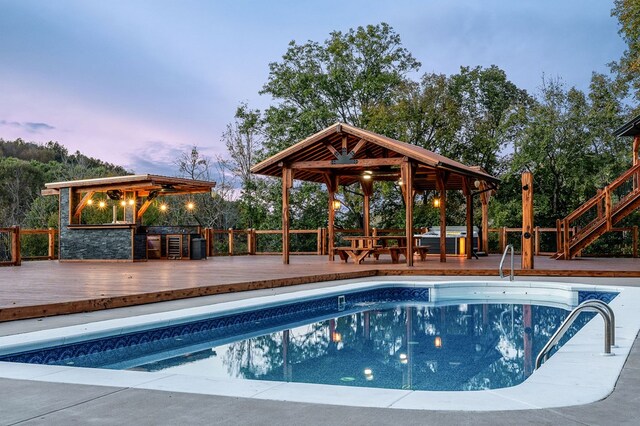 pool at dusk with a gazebo, exterior kitchen, an outdoor bar, a wooden deck, and a fenced in pool