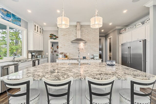 kitchen featuring an island with sink, wall chimney range hood, stainless steel appliances, and decorative light fixtures