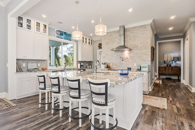 kitchen featuring wall chimney range hood, a center island with sink, glass insert cabinets, and white cabinets