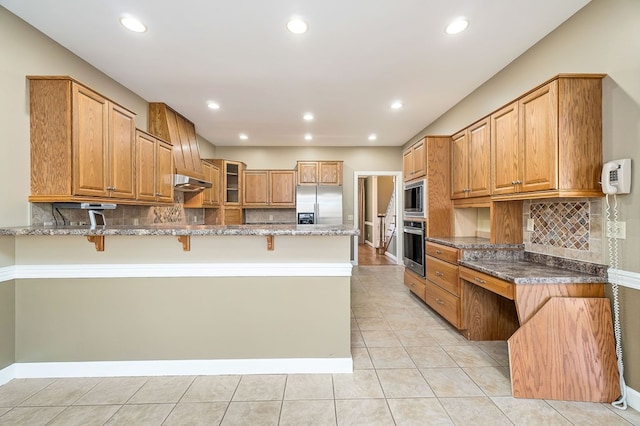 kitchen with stainless steel appliances, dark stone counters, brown cabinetry, a kitchen bar, and glass insert cabinets