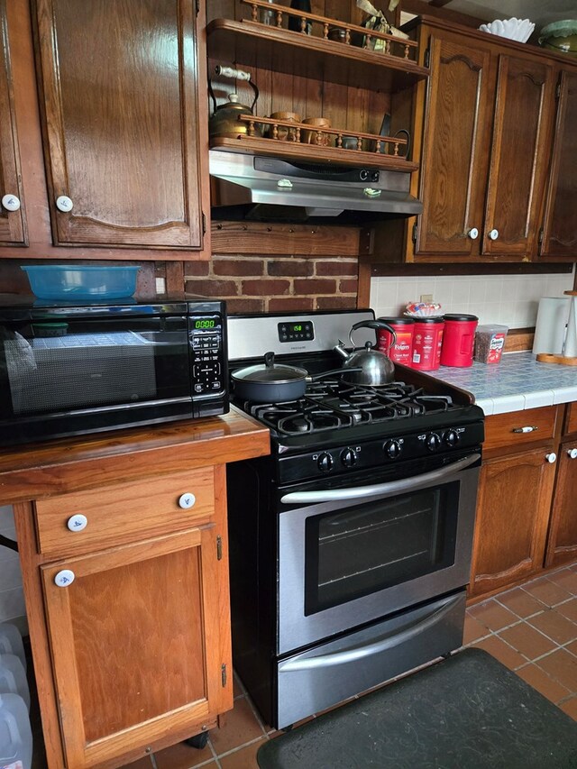 kitchen featuring stainless steel gas range oven, under cabinet range hood, dark tile patterned flooring, brown cabinets, and tasteful backsplash