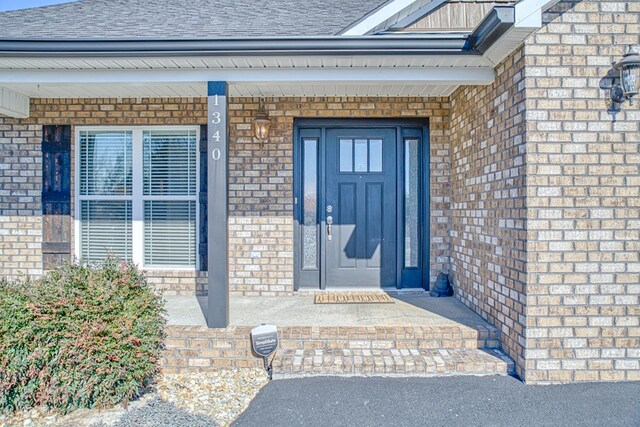 doorway to property with covered porch, brick siding, and roof with shingles