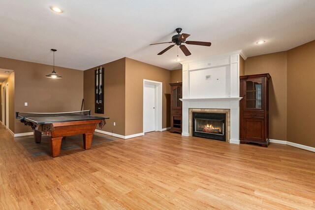 game room featuring light wood-style floors, a warm lit fireplace, baseboards, and a ceiling fan