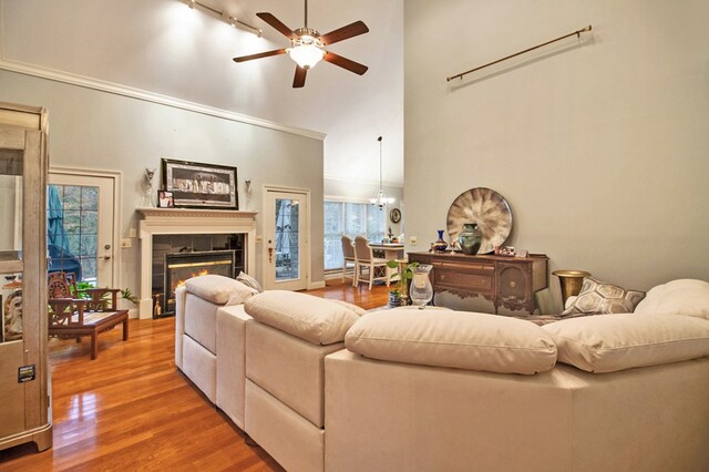 living room featuring a tiled fireplace, ornamental molding, wood finished floors, a high ceiling, and ceiling fan with notable chandelier