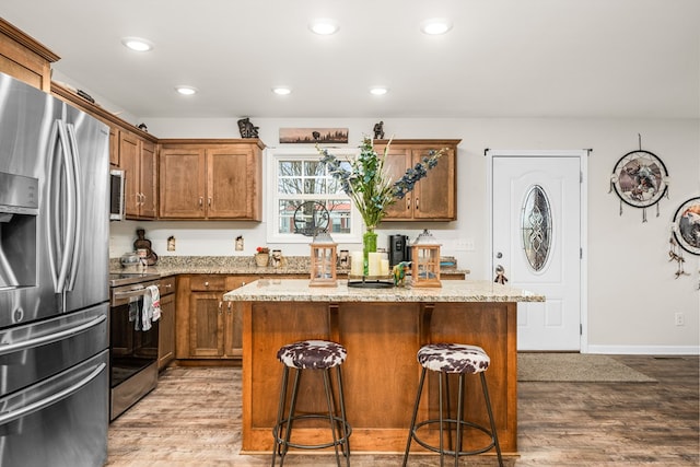 kitchen with stainless steel appliances, light stone countertops, brown cabinetry, a kitchen island, and wood finished floors