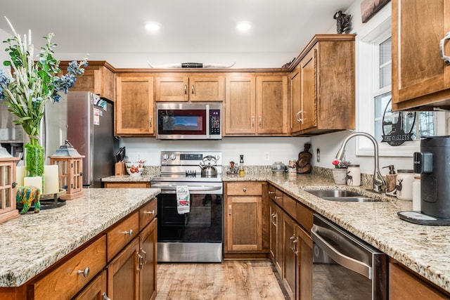 kitchen with brown cabinetry, light wood-style flooring, light stone counters, appliances with stainless steel finishes, and a sink