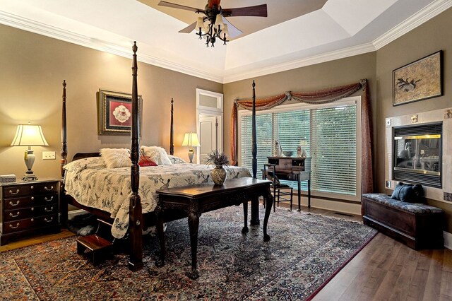 bedroom featuring a tray ceiling, crown molding, dark wood finished floors, a ceiling fan, and a glass covered fireplace