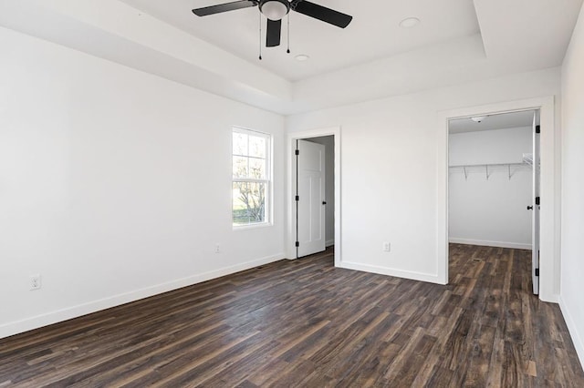 unfurnished bedroom with dark wood-type flooring, a tray ceiling, a walk in closet, and baseboards