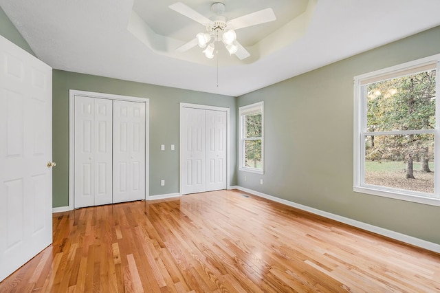 unfurnished bedroom with light wood finished floors, baseboards, a tray ceiling, and multiple closets