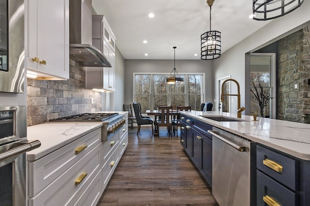 kitchen featuring decorative light fixtures, appliances with stainless steel finishes, white cabinetry, a sink, and wall chimney exhaust hood