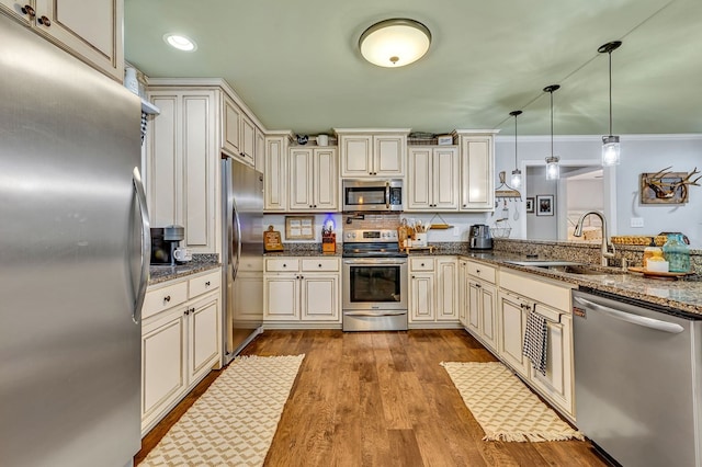 kitchen with cream cabinets, stainless steel appliances, a sink, hanging light fixtures, and dark stone countertops