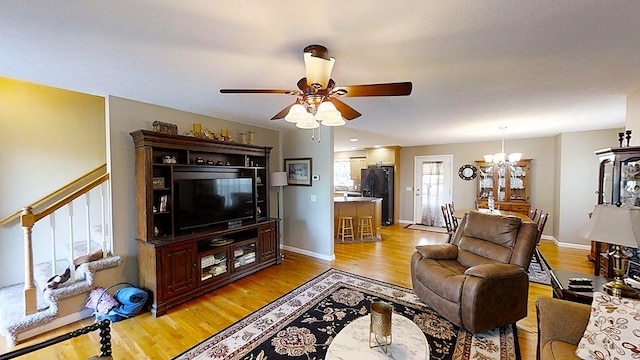 living room featuring light wood-style flooring, stairway, baseboards, and ceiling fan with notable chandelier