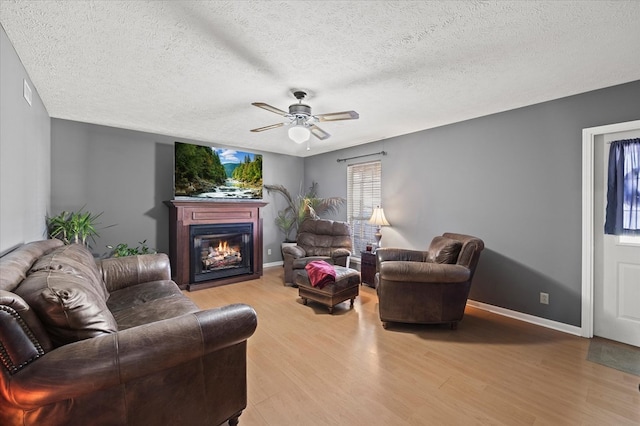 living room featuring a ceiling fan, baseboards, a textured ceiling, a glass covered fireplace, and light wood-type flooring