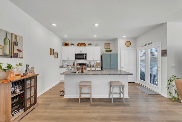 kitchen with light wood finished floors, stainless steel appliances, stone countertops, white cabinets, and a kitchen island with sink