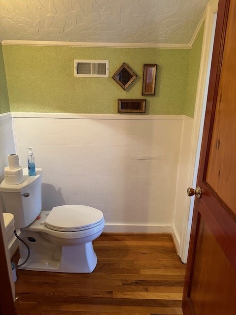 half bath featuring toilet, a wainscoted wall, wood finished floors, visible vents, and ornamental molding