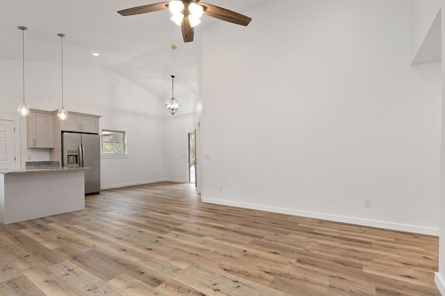 unfurnished living room with ceiling fan with notable chandelier, high vaulted ceiling, light wood finished floors, and baseboards