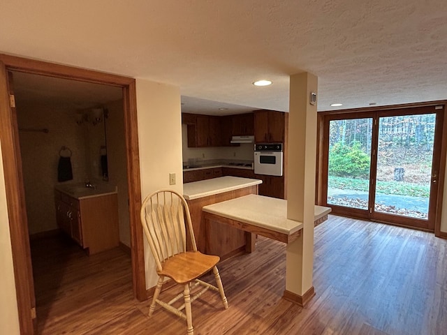 kitchen featuring light countertops, wall oven, a peninsula, wood finished floors, and under cabinet range hood