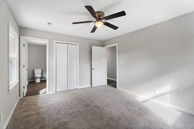 unfurnished bedroom with a closet, dark carpet, a ceiling fan, ensuite bath, and baseboards