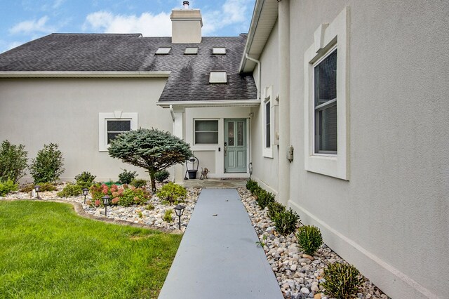 view of exterior entry with a shingled roof, a lawn, a chimney, and stucco siding