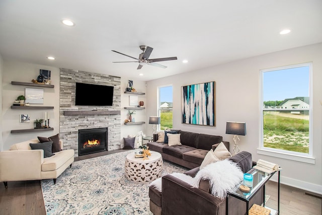 living room featuring a stone fireplace, ceiling fan, dark wood-type flooring, and a healthy amount of sunlight