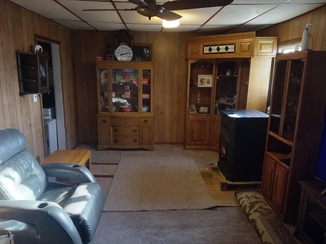 living room featuring a wood stove, ceiling fan, a paneled ceiling, and wood walls