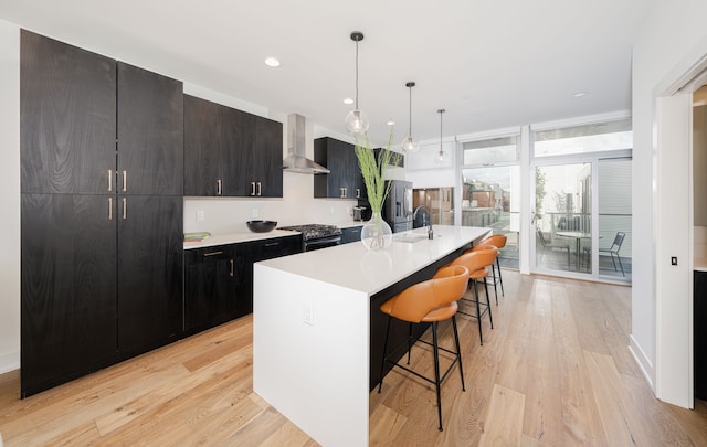 kitchen with a kitchen breakfast bar, wall chimney exhaust hood, stainless steel appliances, light hardwood / wood-style flooring, and an island with sink