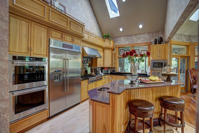 kitchen with built in appliances, an island with sink, a skylight, and wall chimney exhaust hood