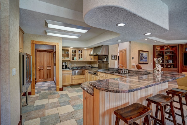 kitchen featuring light brown cabinets, wall chimney exhaust hood, a kitchen breakfast bar, kitchen peninsula, and a textured ceiling