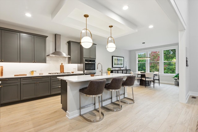 kitchen featuring a center island with sink, pendant lighting, wall chimney range hood, and light wood-type flooring