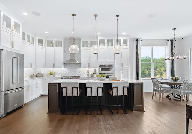 kitchen featuring wall chimney exhaust hood, stainless steel appliances, dark wood-type flooring, decorative light fixtures, and a center island with sink