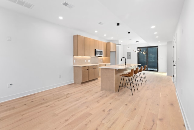 kitchen with backsplash, light hardwood / wood-style floors, decorative light fixtures, light brown cabinetry, and a center island with sink