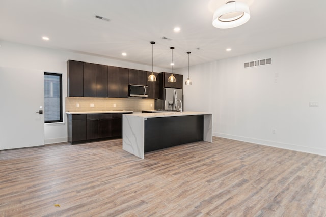 kitchen featuring tasteful backsplash, pendant lighting, a kitchen island with sink, appliances with stainless steel finishes, and light wood-type flooring