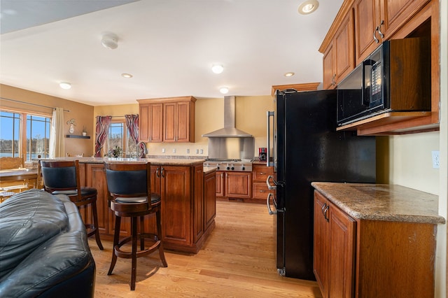 kitchen with a center island, wall chimney range hood, a kitchen breakfast bar, black fridge, and light hardwood / wood-style flooring