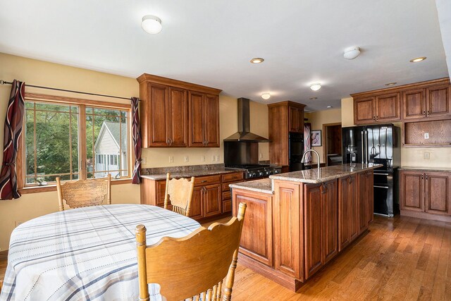 kitchen featuring dark stone counters, wall chimney exhaust hood, an island with sink, light hardwood / wood-style floors, and black fridge with ice dispenser