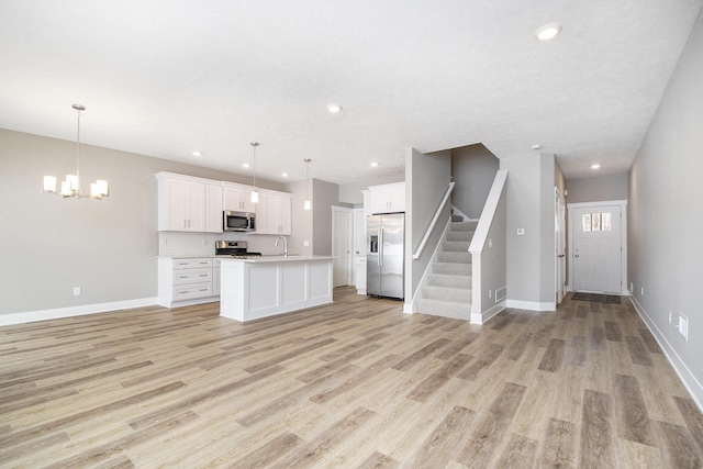 kitchen with appliances with stainless steel finishes, an island with sink, white cabinets, hanging light fixtures, and light hardwood / wood-style flooring