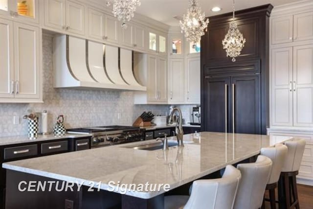 kitchen with white cabinetry, hanging light fixtures, paneled built in fridge, stainless steel range oven, and a kitchen island with sink