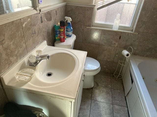 bathroom featuring tile patterned floors, a tub to relax in, toilet, vanity, and tile walls