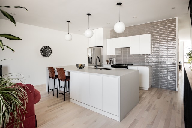 kitchen with stainless steel refrigerator with ice dispenser, a center island with sink, light hardwood / wood-style flooring, white cabinetry, and hanging light fixtures