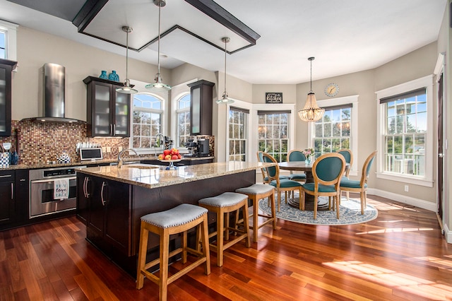 3975 Harbor Breeze Dr, Norton Shores, MI 49441 - kitchen featuring stainless steel oven, wall chimney exhaust hood, dark wood finished floors, glass insert cabinets, and a kitchen island with sink