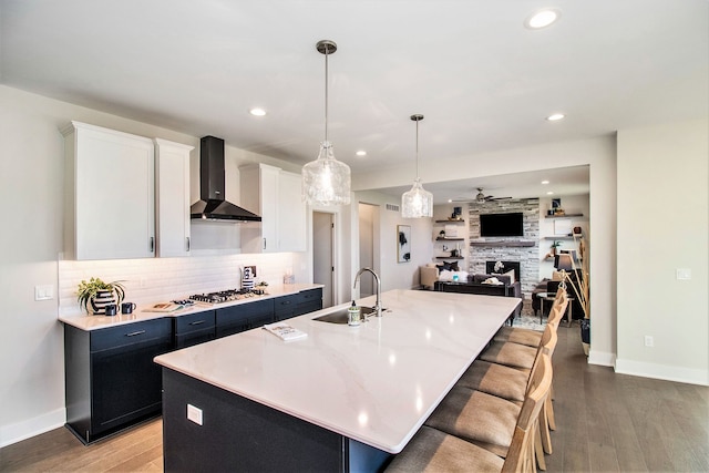 kitchen with wall chimney range hood, sink, an island with sink, decorative light fixtures, and white cabinetry