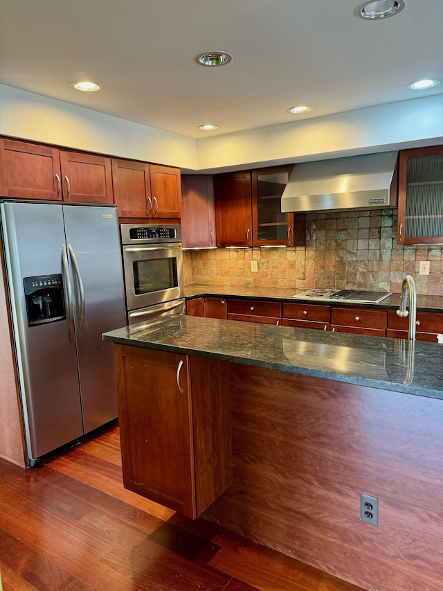 kitchen with wall chimney exhaust hood, stainless steel appliances, dark hardwood / wood-style flooring, backsplash, and dark stone countertops